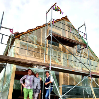 Bild einer LORENZ-Referenz mit dem Bauherrenpaar und der Planerin vor einem albgeplanten Strohbau am gerüsteten Haus mit Richtfestkranz